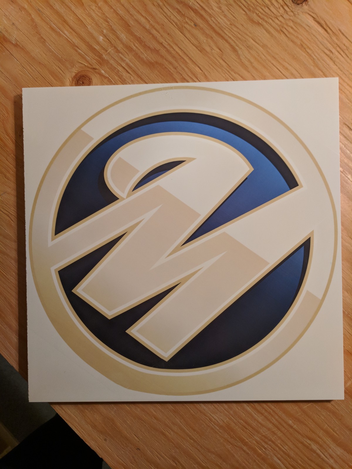 Single custom floor tile with a blue, black and beige circular logo, shown on a wooden worktop before installation.